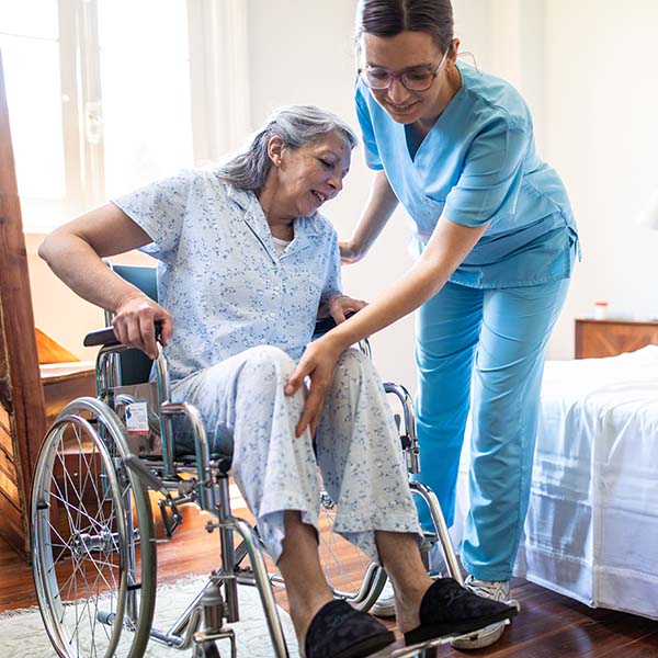 Female nurse helping senior woman to get up from wheelchair in bedroom